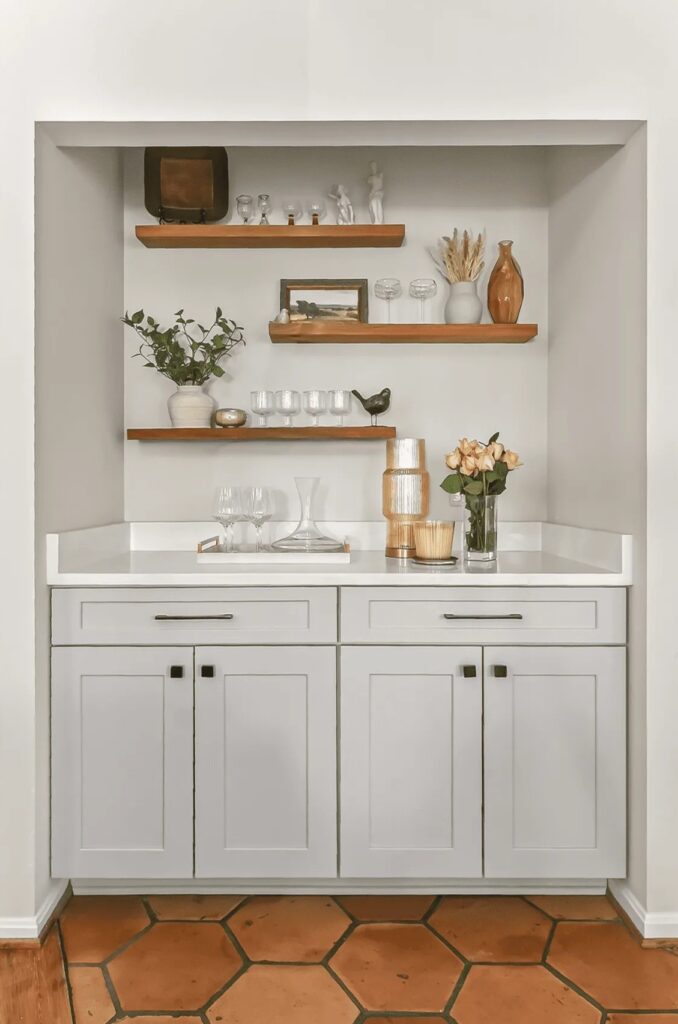 Stylish home bar area featuring open shelves, decorative items, and a white countertop with glassware and flowers.